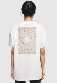 Hvid oversized t-shirt med et beige puslespilgrafik på bagsiden, med et enkelt hvidt puslespilsbrik integreret i designet.