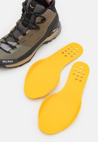 Scarponcino da trekking alto in marrone e grigio con lacci neri, accanto a due solette gialle, con perforazioni e texture liscia.