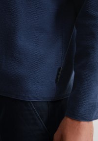 Granatowy, teksturowany sweter z ribbowym dołem i mankietami, z małą metką po boku. Zbliżenie na rękaw i korpus.