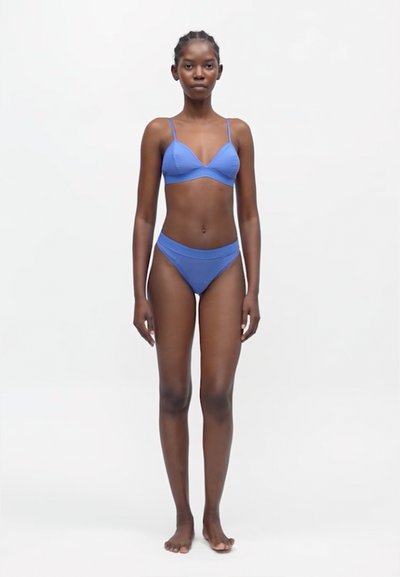 Γυναίκα στέκεται ξυπόλυτη σε λευκό φόντο φορώντας μπλε σουτιέν και αντίστοιχα εσώρουχα με ψηλή μέση.