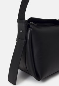 Sac à main en cuir noir avec une large bandoulière réglable et une fermeture éclair visible sur le dessus, posé sur un fond blanc.
