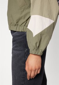 Hand resting by side wearing olive-green jacket with white elbow patch and elastic cuff, paired with dark gray denim jeans.
