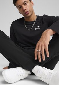 Sort langærmet skjorte med Puma-logo, sorte bukser og hvide sneakers. Sølv kædehalskæde og ring på hånden. Afslappet siddende stilling.