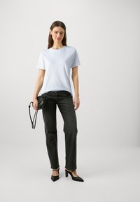 Joven mujer que lleva una camiseta blanca, jeans negros, tacones negros y sostiene un pequeño bolso negro frente a un fondo liso.