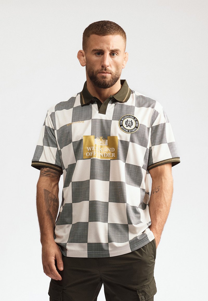 Homme aux cheveux courts et barbe, portant un polo à carreaux noir et blanc avec le logo "Weekend Offender" et un pantalon cargo vert olive.