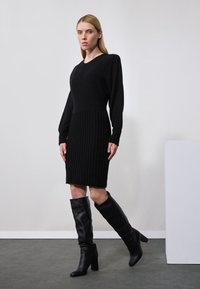 Schwarzes Strickkleid mit V-Ausschnitt, das eine gerippte Textur aufweist. Kombiniert mit hohen schwarzen Lederstiefeln, vor einem neutralen Hintergrund.