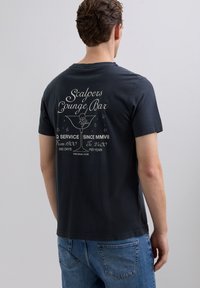 Hombre con camiseta oscura con una impresión en la espalda que muestra "Scalpers Lounge Bar", un vaso de cóctel con una calavera, horas de servicio y el texto "The Skull Club".