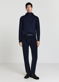 Marineblaue Strickpullover mit Kragen und drei Knöpfen, kombiniert mit passenden plissierten Hosen und schwarzen Schnürschuhen, vor einem schlichten Hintergrund.