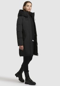 Junge Frau trägt einen langen schwarzen Daunenmantel mit Kapuze, schwarze Leggings und schwarze klobige Stiefel, die vor einem schlichten hellen Hintergrund geht.