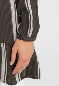 Brązowa bluzka o teksturze z białymi poziomymi paskami, z luźnymi rękawami i falbaniaste wykończeniami, pokazana od pasa w górę, z ręką na materiale.