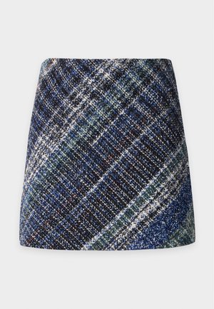 Donna con lunghi capelli biondi che indossa un top senza maniche con motivo, una minigonna scozzese blu e sandali con tacco alto, in piedi su uno sfondo neutro.