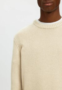Stickad tröja i beige med ett strukturerat mönster, har en rund halsringning och ribbad nederkant. Tyget verkar mjukt och bekvämt.