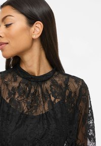 Femme portant un haut en dentelle noire à motifs floraux avec un col montant, des manches transparentes, une petite boucle d'oreille en perle et un eye-liner bleu, regardant sur le côté.