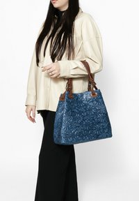 Sac à main bleu à relief avec des motifs floraux, poignées en cuir marron et une forme structurée. Porté sur le côté contre un fond neutre.