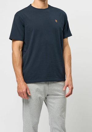 Mand iført en navyblå kortærmet t-shirt med en lille broderet logo og lysegrå bukser mod en ensfarvet baggrund.