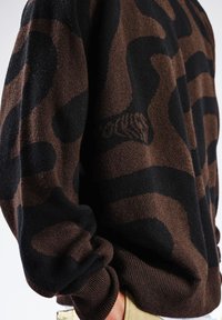 Pull à motifs marron et noir, avec une texture douce, des poignets retroussés et une coupe décontractée. Design abstrait distinctif sur tout le tissu.