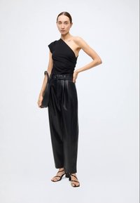 Schwarzes asymmetrisches Oberteil mit einer Schulter, kombiniert mit hoch taillierter schwarzer Lederhose. Das Modell hält eine kleine schwarze Handtasche. Schwarze Riemchensandalen.