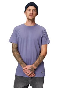 Lilla bomulds-T-shirt med korte ærmer, rund hals og logomærke ved kanten. Modellen har tatoveringer og bærer en marinblå hue.
