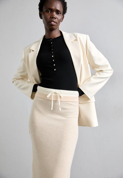 Cremfarbener Blazer über einem schwarzen Ripp-Top mit Knopfdetails, kombiniert mit einem hellbeigen Rock mit elastischem Bund und Zugband.