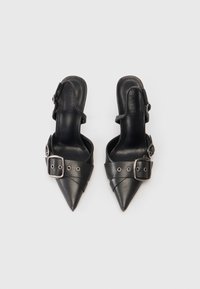 O pereche de pantofi slingback negri, cu vârful ascuțit, curele lata și detalii cu catarame argintii, pe un fundal alb.