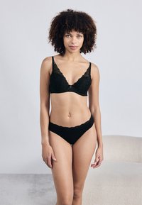 Μαύρο σουτιέν από δαντέλα με βυθισμένο ντεκολτέ και ρυθμιζόμενες τιράντες. Το αντίστοιχο μαύρο μπικίνι από δαντέλα με κομμένη άκρη ολοκληρώνει το σετ.