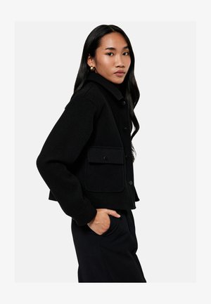 Vrouw met lang donker haar die een zwarte, geknoopte jas met grote zakken draagt, poseert met één hand in de zak tegen een effen achtergrond.