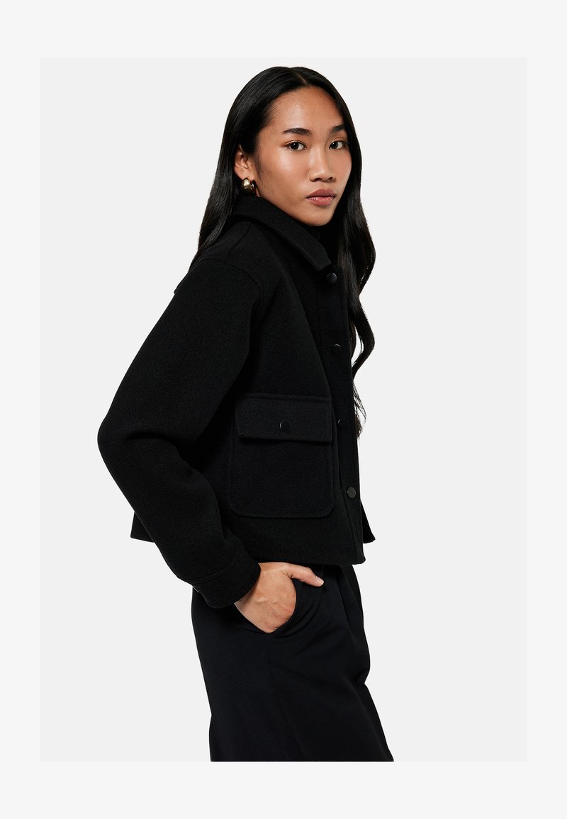 Kobieta z długimi ciemnymi włosami, ubrana w czarną zapinaną na guziki kurtkę z dużymi kieszeniami, pozująca z jedną ręką w kieszeni na jednolitym tle.