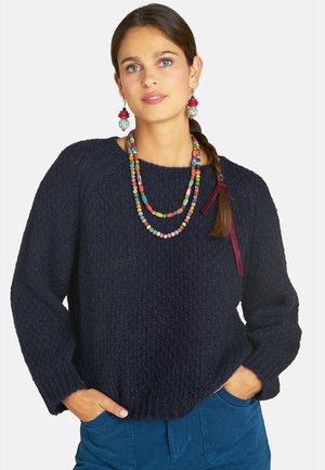 Frau mit langem geflochtenem Haar, die einen dunkel texturierten Pullover, eine bunte Perlenkette, passende Ohrringe und blaue Hose trägt, mit Händen in den Taschen.