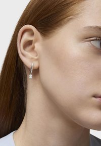 Orecchino a cerchio in argento con un piccolo ciondolo bianco appeso nella parte inferiore. L'orecchino è indossato all'orecchio di una modella su una pelle liscia.