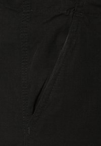 Svart tyg med en subtil textur och en sidoficka. Nära sydda sömmar framhäver plaggets konstruktion.