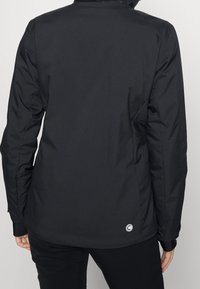 Person, der eine taillierte schwarze Jacke trägt, mit einem kleinen runden Logo in der Nähe des unteren Saums, von hinten gezeigt.