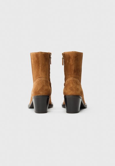 O pereche de boots din suede maro, până la glezne, cu toc pătrat, închidere cu fermoar la spate și talpă din lemn neted, pe un fundal alb.