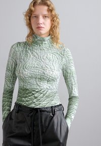 Long-sleeve turtleneck top in textured light green fabric with quilted details, paired with high-waisted black faux leather pants.