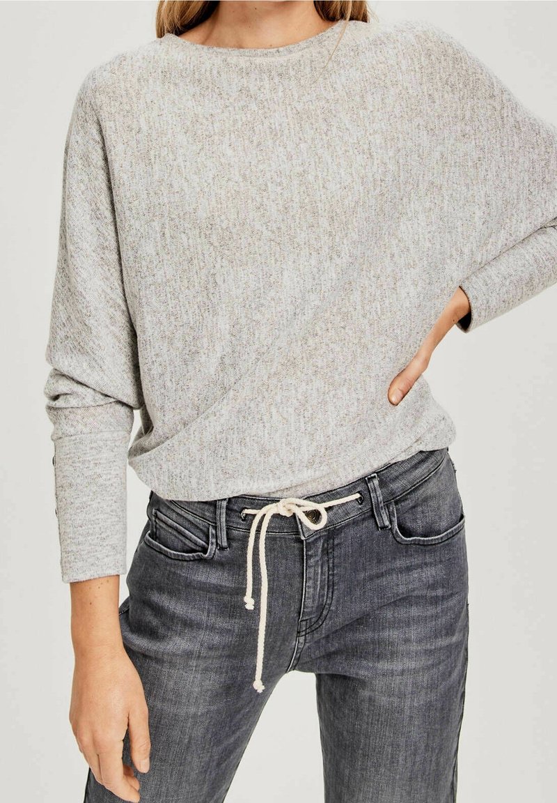 Jasnoszary, teksturowany sweter z okrągłym dekoltem i opuszczanymi ramionami, zestawiony z ciemnoszarymi dżinsami z wiązaniem w pasie.