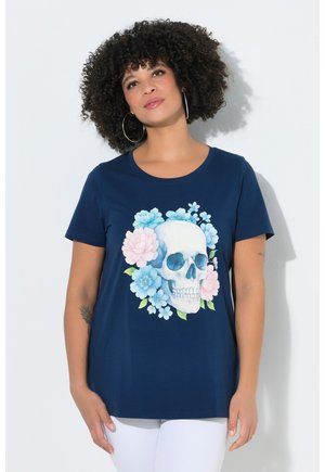 Frau mit lockigem Haar trägt ein marineblaues T-Shirt mit einem weißen Totenkopf, umgeben von blauen und pinken Blumen, kombiniert mit weißen Hosen.