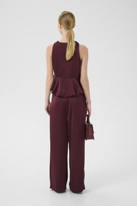 Žena s blond copom, oblečená v bezrukávovom bordovom peplum topu a širokých nohaviciach, drží malú štruktúrovanú bordovú kabelku, pohľad zozadu.