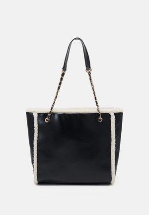 Sac cabas en cuir noir avec garniture en peau lainée blanche et anses chaîne dorées entremêlées de cuir noir sur fond uni.