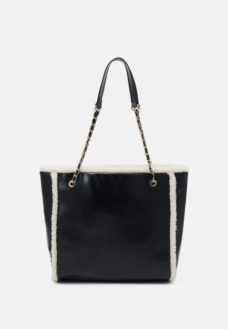 Sac cabas en cuir noir avec garniture en peau lainée blanche et anses chaîne dorées entremêlées de cuir noir sur fond uni.