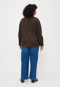 Giacca in suede marrone con taglio dritto, indossata sopra jeans a gamba larga blu. Il retro presenta un semplice design con cuciture e nessun hardware visibile.