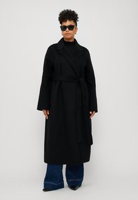 Schwarzer Wollmantel mit langen Ärmeln und Gürtel, ausgestattet mit einem abgerundeten Kragen. Kombiniert mit dunklen Schlagjeans und Sonnenbrille. Glatte Textur.