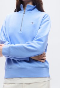 Personne portant un sweat-shirt bleu clair à fermeture éclair quart avec un petit logo sur la poitrine et une chemise blanche en dessous, bras croisés à la taille.