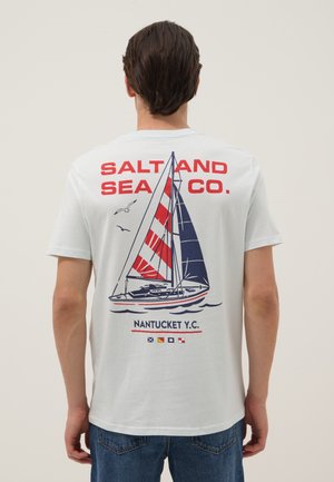 Man wearing white t-shirt with red and navy sailboat graphic and "Salt and Sea Co. Nantucket Y.C." text on the back.