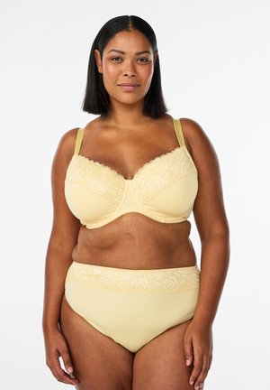 Woman wearing a matching pale yellow lace bra and high-waisted underwear, standing against a plain white background.