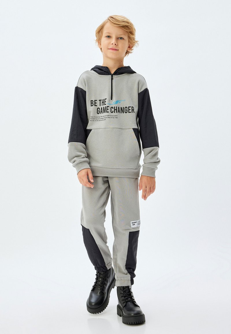 Grå och svart hoodie med dragkedja och grafisk text, kombinerad med matchande joggers med svarta accenter och elastiska manschetter, bärs med svarta stövlar.