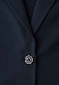 Gros plan sur le tissu d'un blazer bleu marine foncé avec un seul bouton noir portant l'inscription "Street One" et des coutures visibles sur le revers.