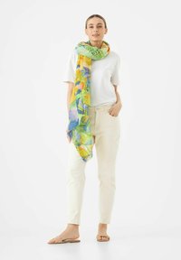 Foulard floral en vert, jaune et bleu, en tissu léger, drapé autour du cou sur une chemise blanche et un jean crème.