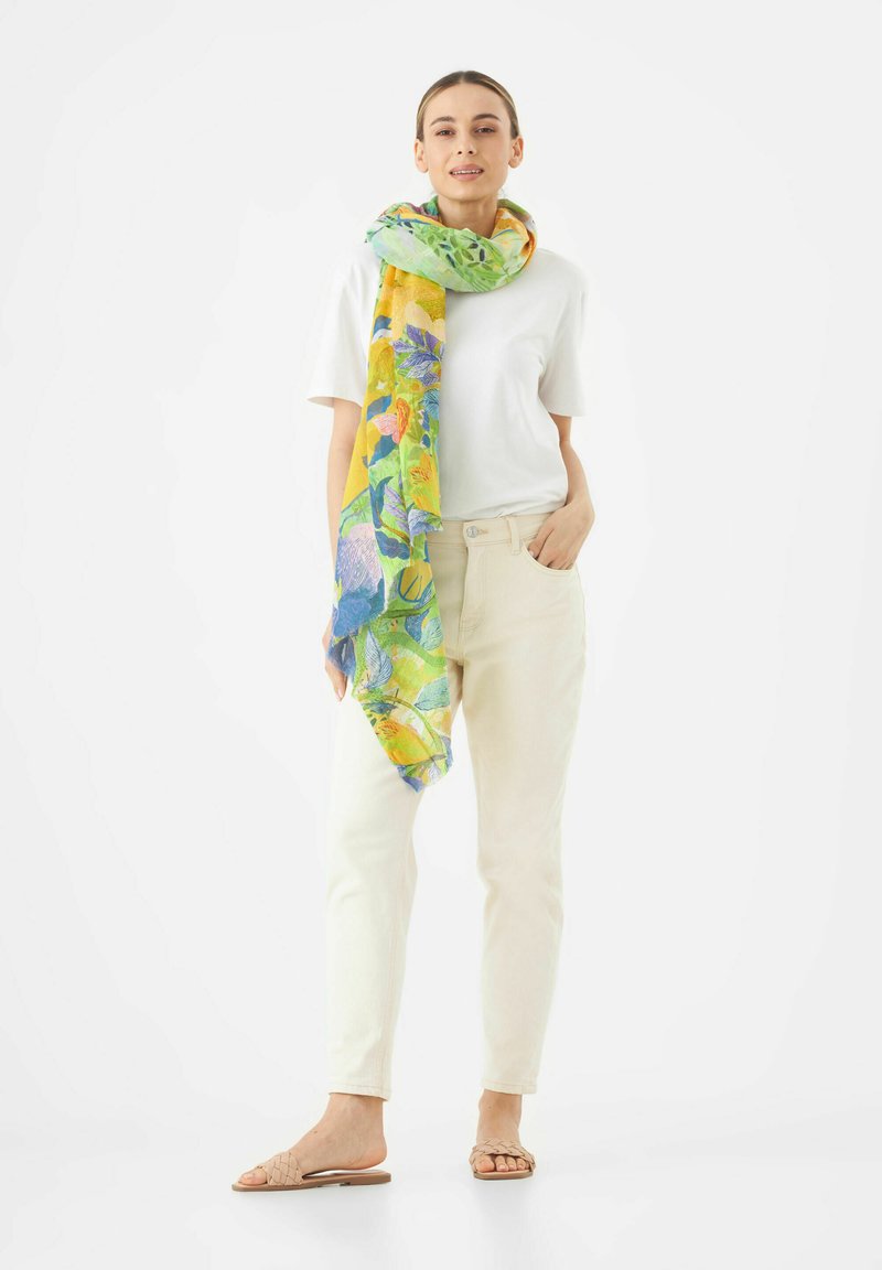Foulard floral en vert, jaune et bleu, en tissu léger, drapé autour du cou sur une chemise blanche et un jean crème.