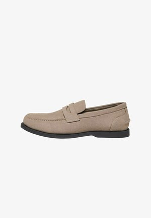 Mocassin beige en daim à enfiler avec semelle noire, bout arrondi et coutures décoratives sur les côtés et le dessus.