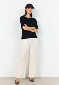 Kvinna står med armarna i kors, iförd svart keps, svart kortärmad topp, vita vida byxor och klackar i zebramönster framför en vit bakgrund.