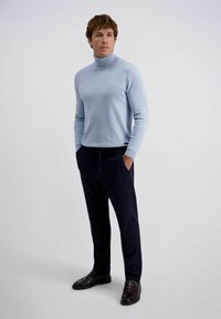 Man die staat met handen in zijn zakken, gekleed in een lichtblauwe coltrui, donkere broek met trekkoord en donkere instappers op een witte achtergrond.